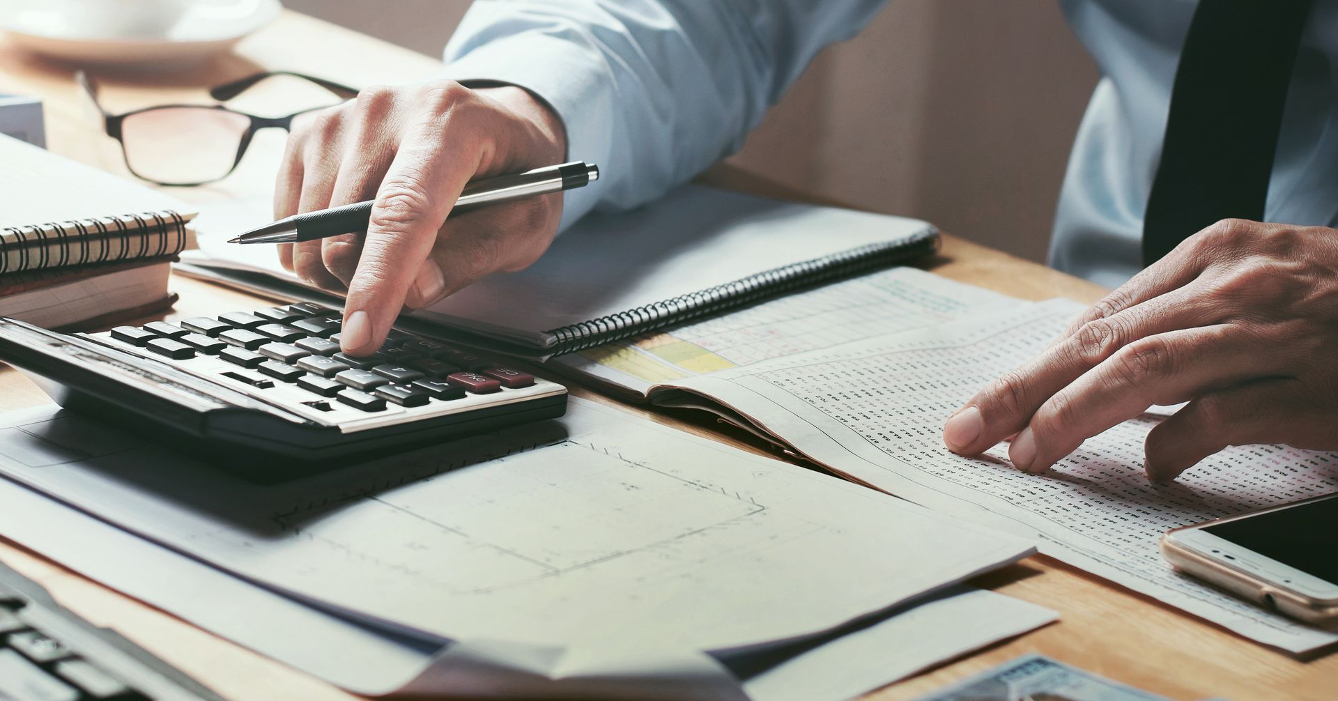 Person arbeitet an einem Schreibtisch mit Taschenrechner, Stift und aufgeschlagenen Finanzdokumenten bzw. Tabellen